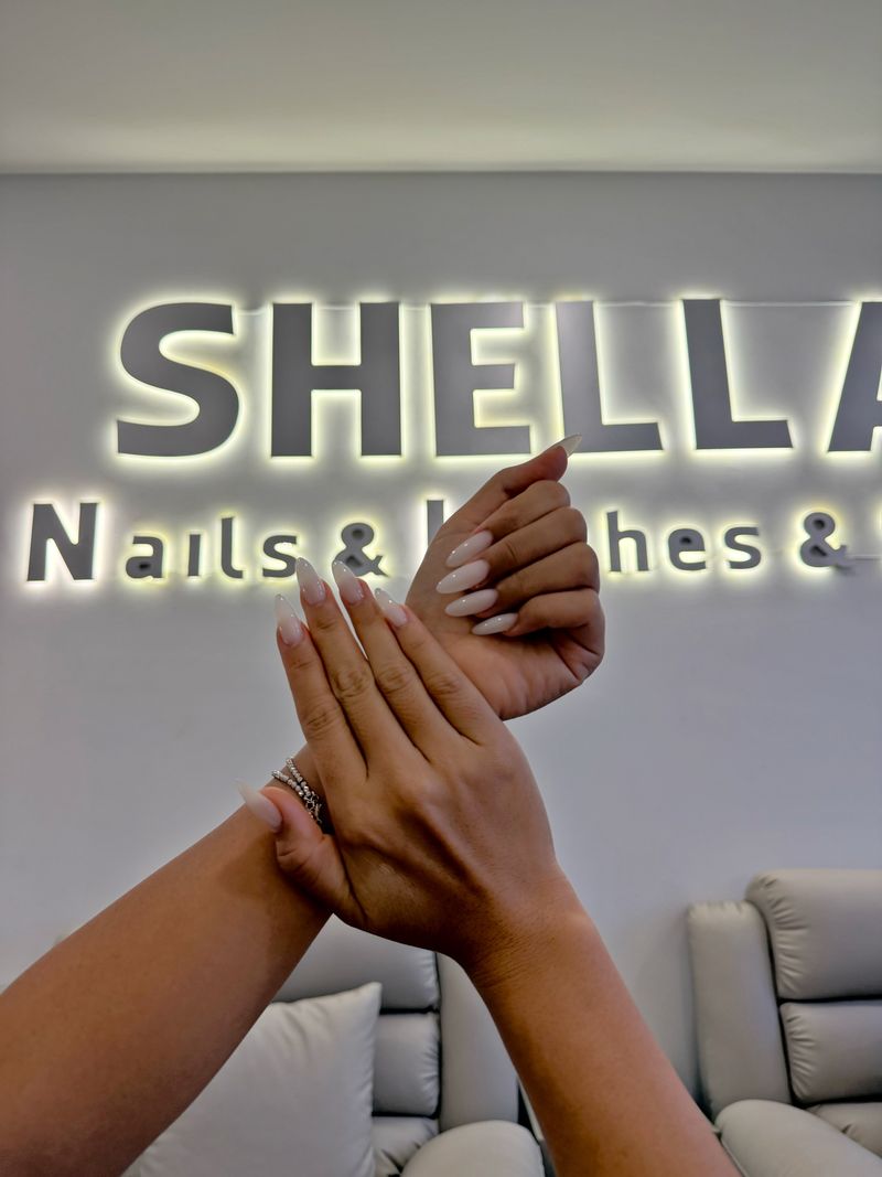 Длинные миндалевидные ногти молочно-белого цвета на фоне вывески Shella Nails & Lashes в Дананге